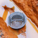 A close-up image shows a round metal drain cover partially submerged in muddy water. The surrounding area consists of orange-brown soil and scattered small stones. The water has a brownish tint, likely from the mud. The drain cover features a grid pattern with vertical and horizontal slots.