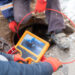 Two workers are seen conducting a sewer inspection. One is holding an orange handheld monitor displaying a live video feed from a camera inserted into a manhole. They are wearing red gloves and protective clothing. The second worker is handling the cable attached to the camera, also wearing protective footwear. Snow is visible on the ground, indicating cold weather conditions.